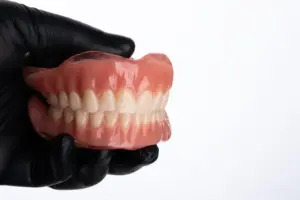 Prótese dentária total (dentadura) superior e inferior sendo segurada por luva preta, demonstrando encaixe e oclusão dos dentes artificiais.