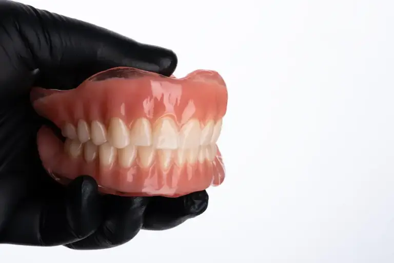 Prótese dentária total (dentadura) superior e inferior sendo segurada por luva preta, demonstrando encaixe e oclusão dos dentes artificiais.